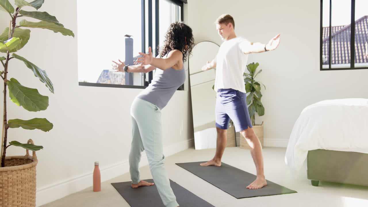 Young multiracial couple practicing yoga together on mats in bright bedroom, at home