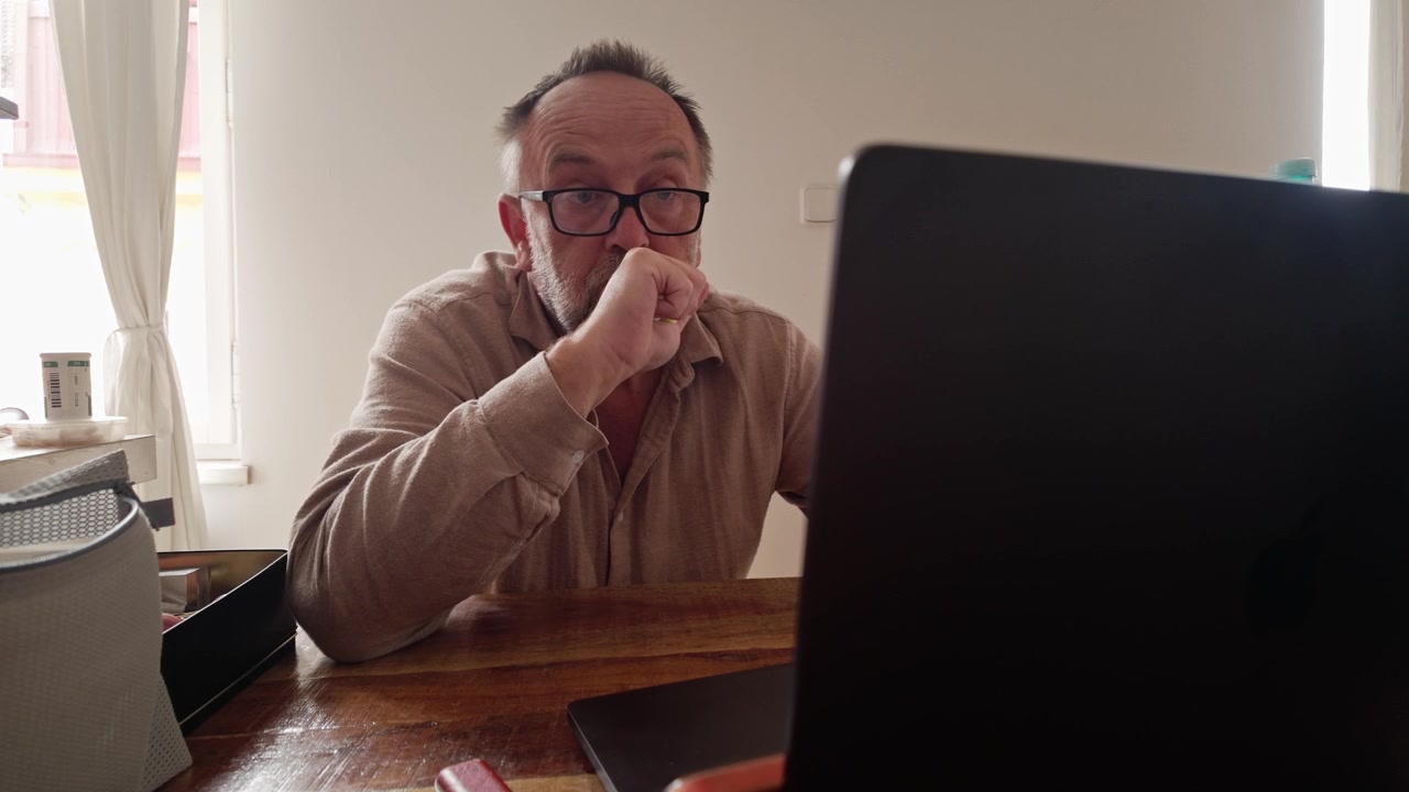 Gestresster Mann drückt einen Stressball, während er zu Hause am Laptop arbeitet, was Müdigkeit und den Druck der Telearbeit zeigt.