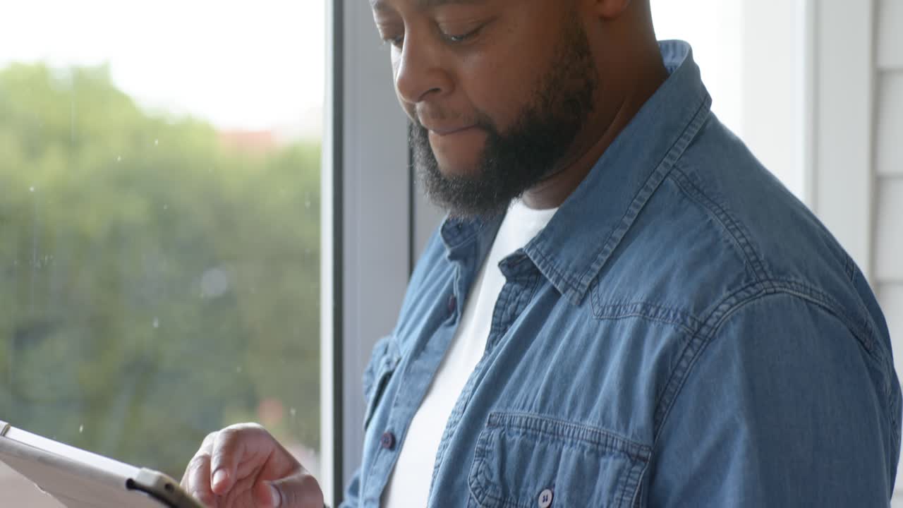 African American man denim-shirt holding tablet and stylus, tapping screen and checking near window