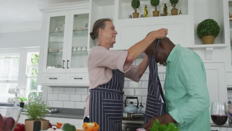 Smiling-senior-diverse-couple-wearing-blue-aprons-and-cooking-in-kitchen