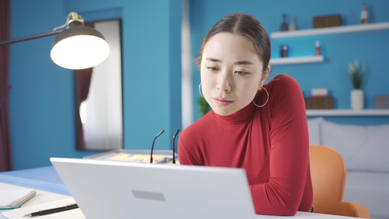 젊은 아시아 여성이 노트북에서 집중적으로 읽고, 안경을 벗고 계속 읽고 있습니다.