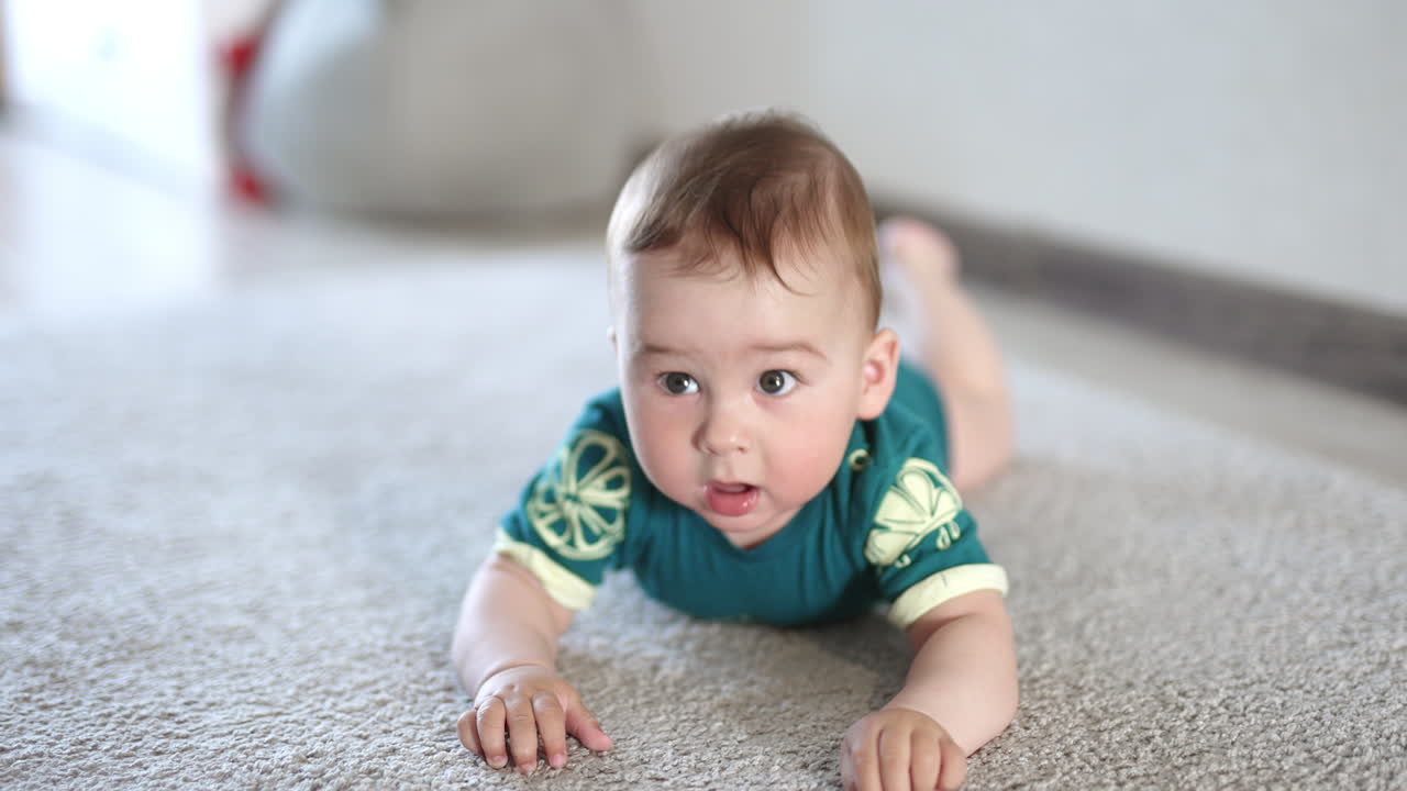 Sweet Caucasian baby boy lies on the carpet banging hands over the floor. Cute infant child holds still a little looking at camera attentively.