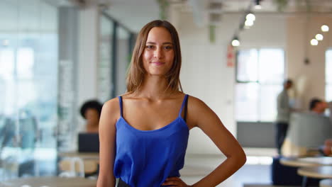 Portrait-Of-Young-Smiling-Businesswoman-Standing-In-Busy-Modern-Open-Plan-Office