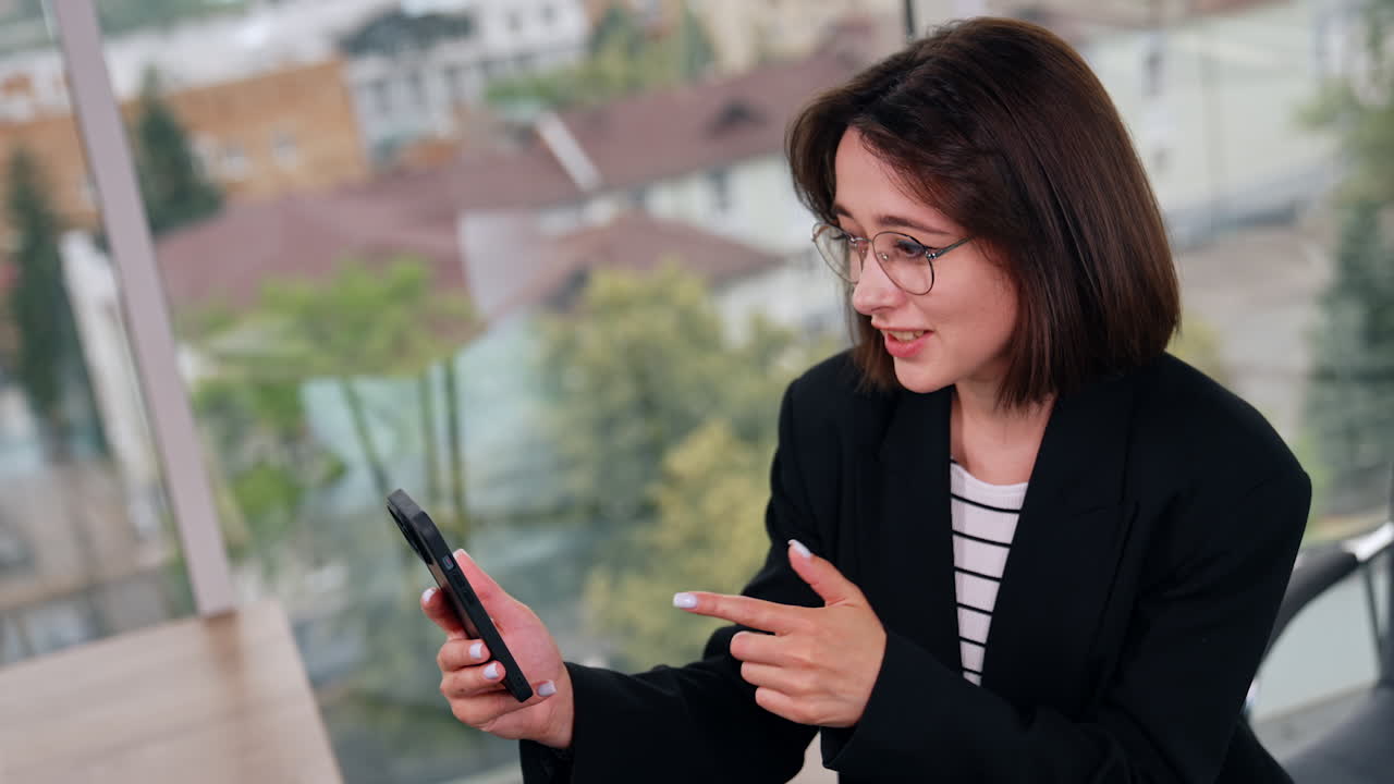 Lady in glasses looks at her smartphone expressing surprise. Top view on the woman having chat online, smiling cutely. Blurred view in the panoramic window at backdrop.