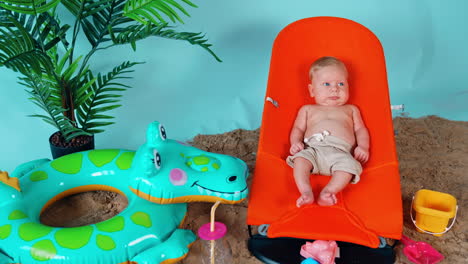 tiny infant blond boy wearing shorts lies in the orange baby chair. little kid on a beach vacation.