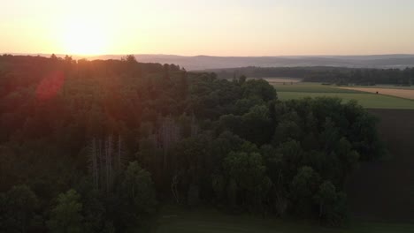 Imágenes-De-Vuelo-Hacia-Atrás-Sobre-Un-Enorme-Bosque-Y-Prados-En-Una-Puesta-De-Sol-Naranja-Dando-Hermosos-Destellos-De-Sol-En-Las-Imágenes-Con-Montañas-En-El-Fondo