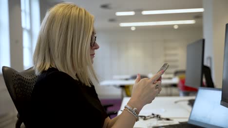 Blonde-Haired-Woman-Sitting-At-Desk-In-Office-Holding-And-Swiping-On-Mobile-Phone