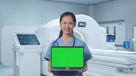 asian female doctor smiling and showing green screen laptop while standing with mri machine