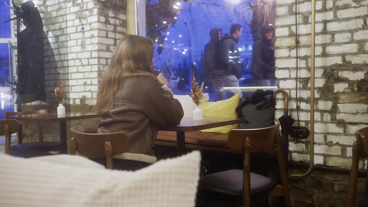 Back shot of woman in brown shearling jacket sitting at cozy cafe sipping coffee, with people walking outside the window. Soft lighting creates a calm and peaceful atmosphere