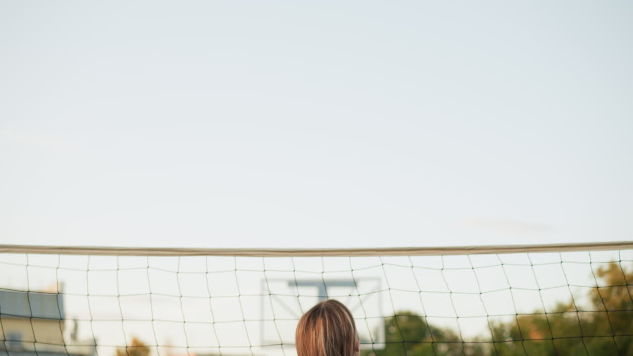 볼리볼을 하는 여자들의 뒷모습: 한 선수가 공을 치기 위해 준비하는 동안, 네트의 반대편에 있는 팀 동료들은 그들과 맞서고, 배경에는 볼리볼 코트와 네트가 있다.