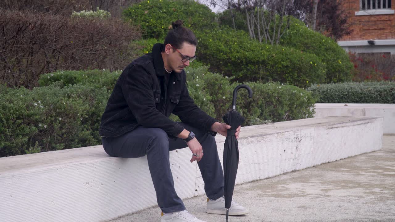 Un giovane, che indossa una giacca nera e occhiali, è seduto su una panchina con un'espressione triste e frustrata.