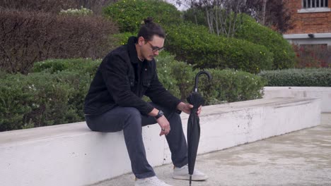 A-young-man,-wearing-a-black-jacket-and-glasses,-sits-on-a-bench-looking-sad-and-frustrated