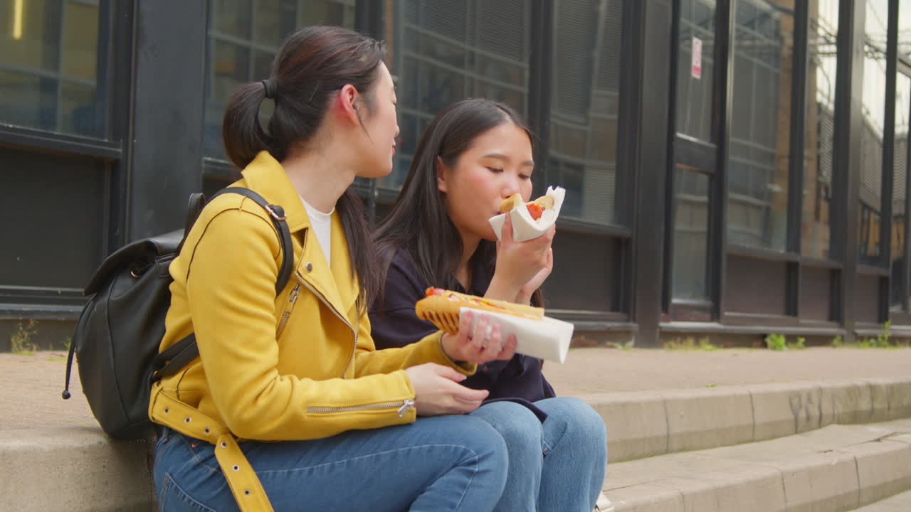 dos jóvenes amigas sentadas en los escalones comiendo perritos calientes comprados en el puesto del mercado de comida callejera