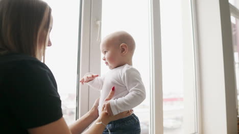 young mother is holding her son's hands while he is walking on the window sill decorated with christmas wreath. happy family kissing