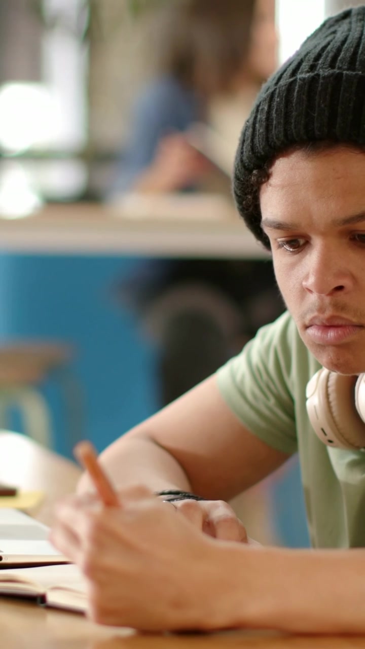 Vertical video: Following adult male studying at cafe, writing with pencil, notebook, beanie
