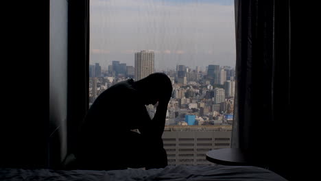 고층 창문 근처에 불안하게 앉아있는 성인 남자의 실루, 배경에 도시 대도시의 파노라마 뷰와 함께 손에 머리를 흔들고