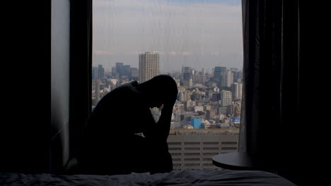 silhouette of adult man anxiously sitting near high rise window, shaking his head in his hands with panoramic view of the city metropolis in background