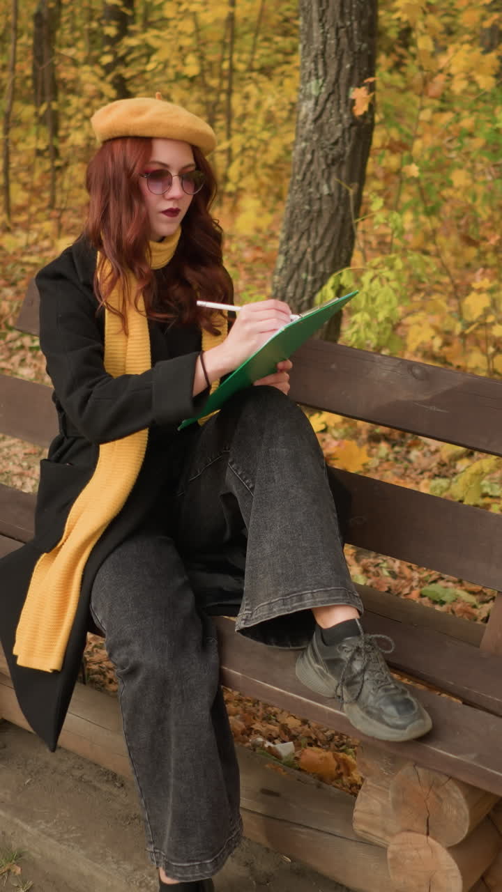 vista aérea de un artista dibujando pensativamente mientras está sentado en un banco de madera en un parque en un entorno tranquilo de otoño, una pierna en el banco, otra en el suelo, rodeado de hojas doradas