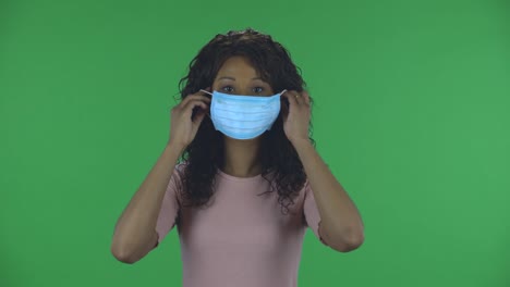 portrait of beautiful african american young woman puts on medical mask. burning brunette with wavy hair in jeans and a beige blouse on a green screen in the studio. health protection corona virus concept