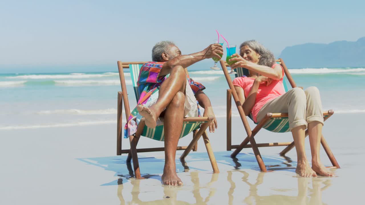 Front view of active senior African American couple toasting drinks on deckchair on the beach 4k
