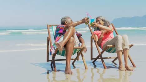 front view of active senior african american couple toasting drinks on deckchair on the beach 4k