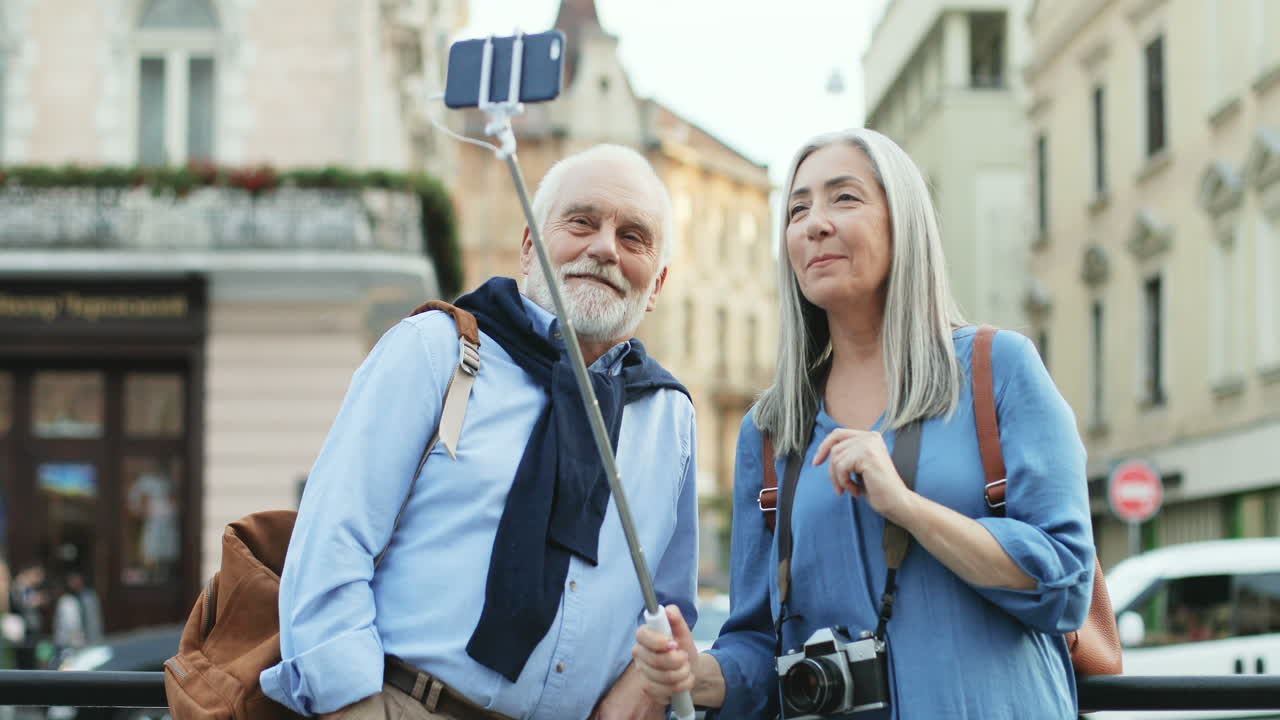 un par de turistas sonrientes parados en el centro de la ciudad con un smartphone en el selfie stick y teniendo un videochat o tomando fotos