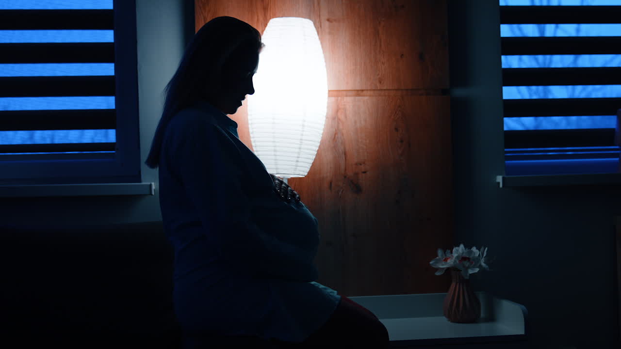 Silhouette of pregnant lady sitting on the bed in dark room. Woman strokes her big belly.