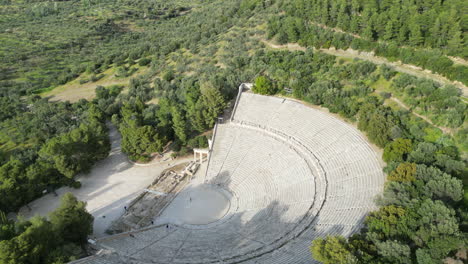 aerial drone shot of the epidaurus theatre in greece, showcasing its grand scale of theatre compared to a person at its center, surrounded by trees