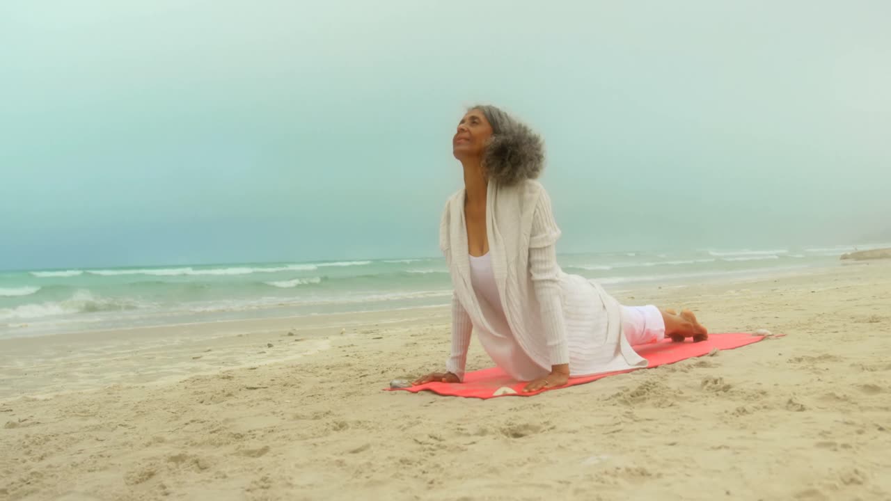 Front view of active senior African American woman doing yoga on exercise mat at the beach 4k