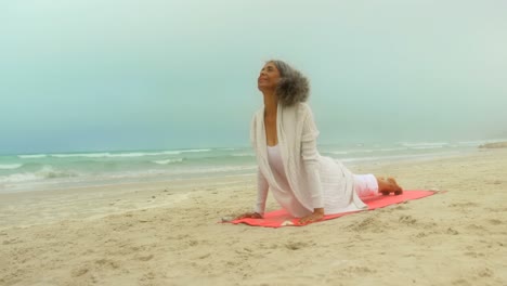 Front-view-of-active-senior-African-American-woman-doing-yoga-on-exercise-mat-at-the-beach-4k