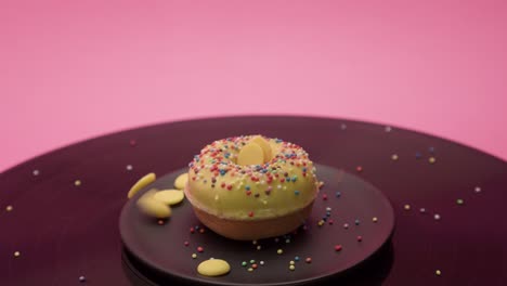 the american donut rolls in circles on a pink and black surface. it is topped with yellow chocolate and garnished with colorful crumbs falling over it