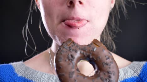 close-up portrait of girl biting and chewing delicious glazed brown donut with chocolate chips on black background.