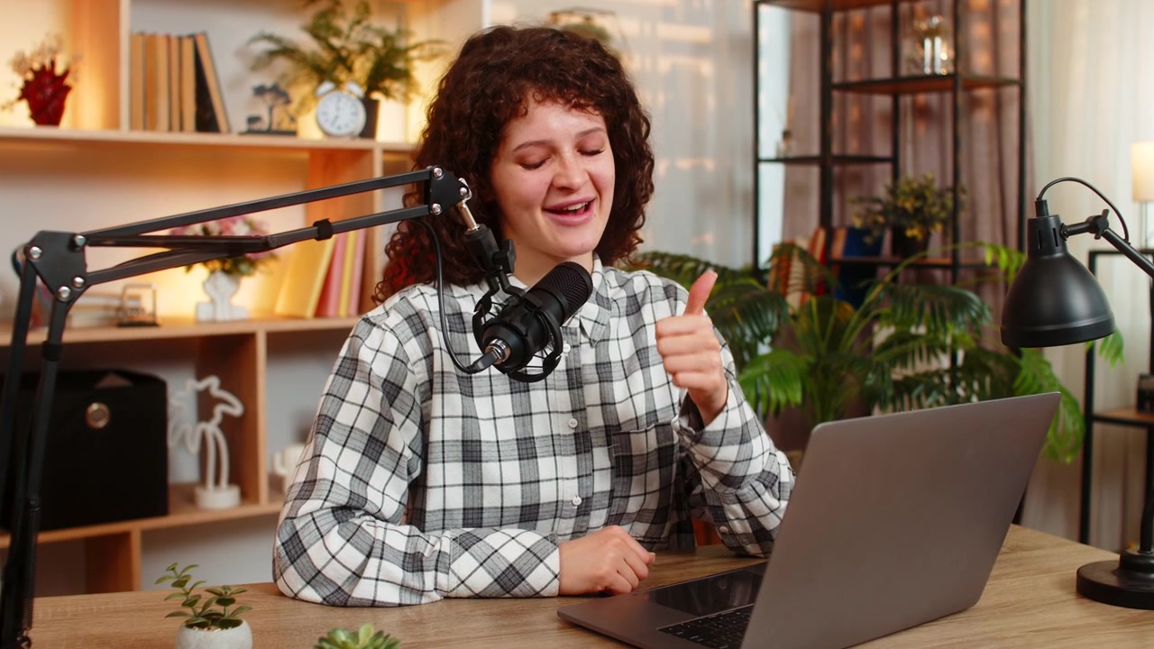Jovem mulher em uma mesa em casa, gravando uma transmissão ao vivo com o microfone do laptop e gesticulando expressivamente.