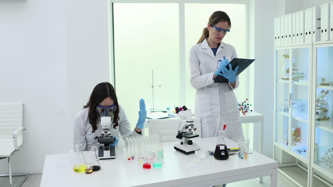 Científico mira por el microscopio mientras un colega escribe un informe en el laboratorio