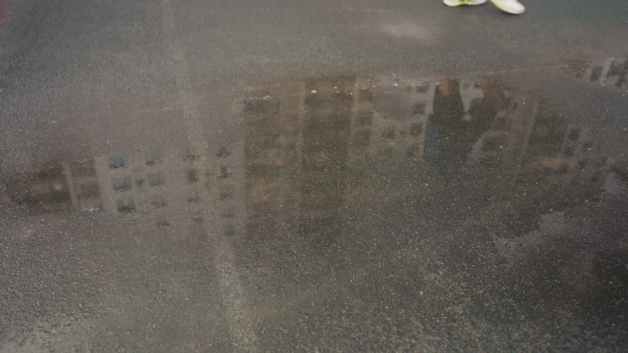 Våd asfaltpyt, der afspejler bygningsfacade og utydelig sko i kanten, let regn, der skaber krusninger, overskyet himmel, der giver grå toner, tekstureret fortov og subtil bysilhuet, rolig regnvejrsdag