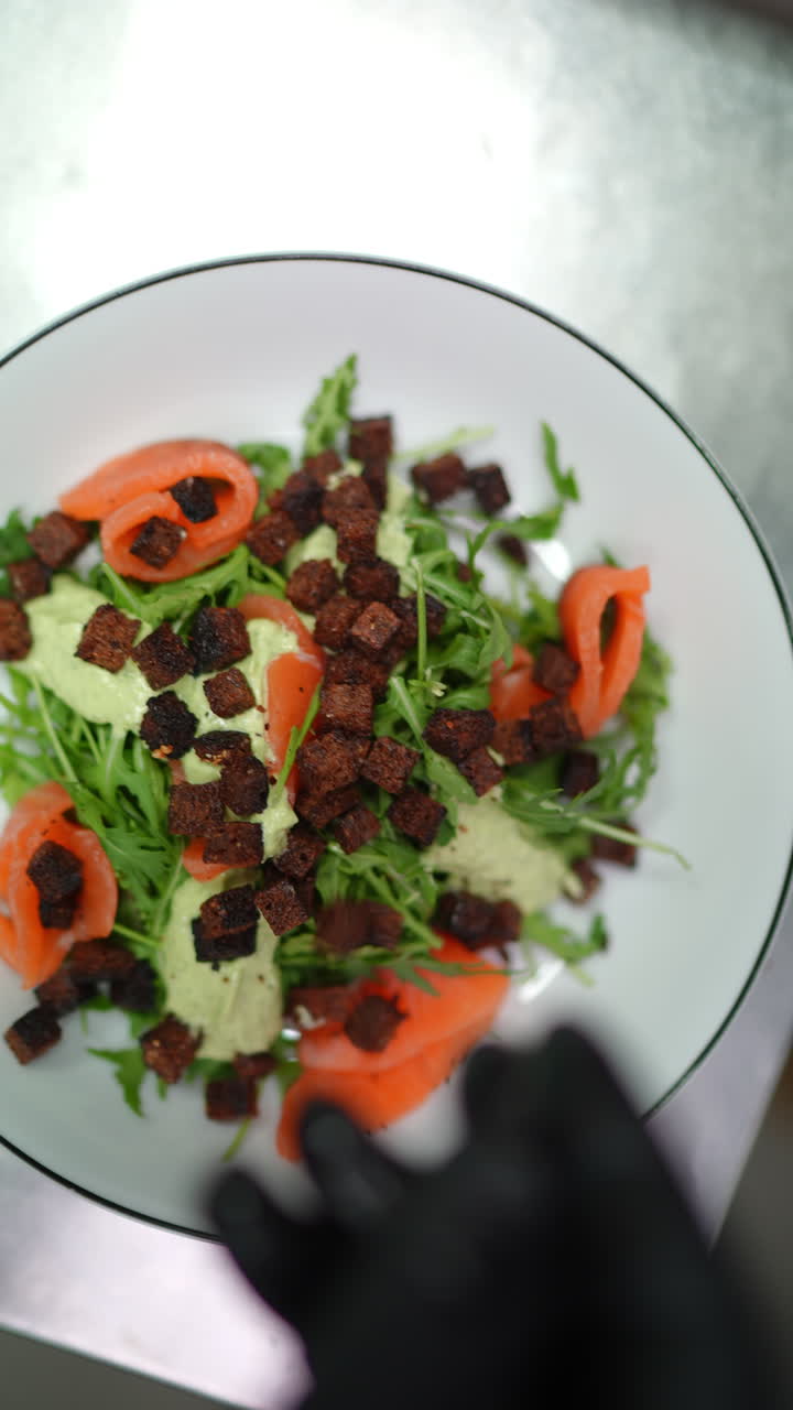 Vertical footage Professional chef wearing black gloves carefully sprinkling crispy croutons over gourmet salmon salad with fresh arugula, adding final elegant garnish on white plate