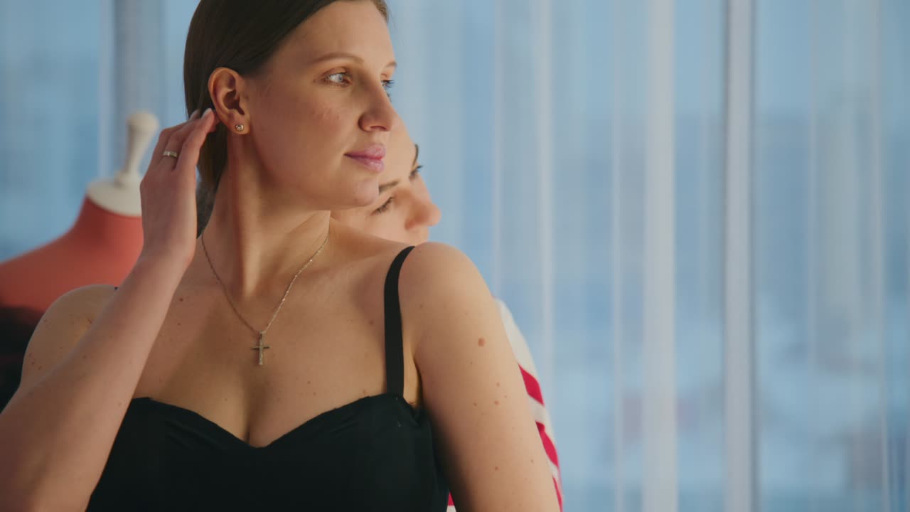 Two women stand side by side, looking out a window. The woman in black appears calm and thoughtful. The woman in red stripes shows concern