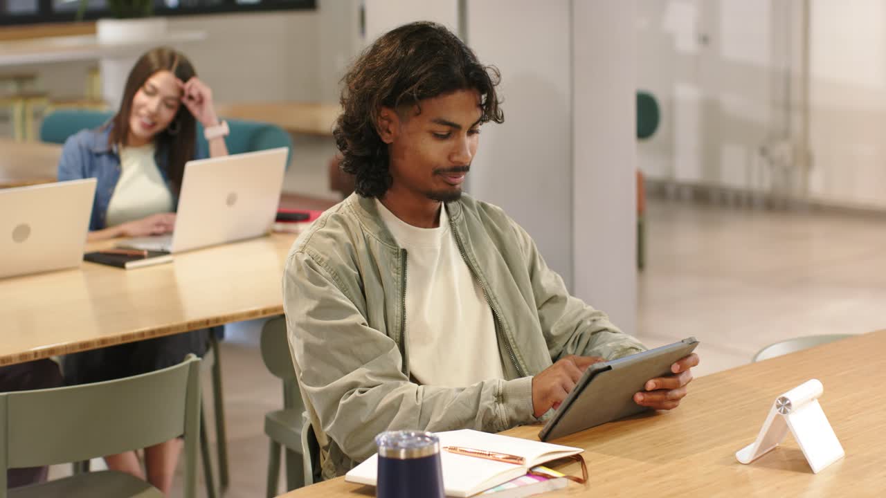 Diverse coworkers sharing table, man in green jacket picking up tablet, tapping through study notes