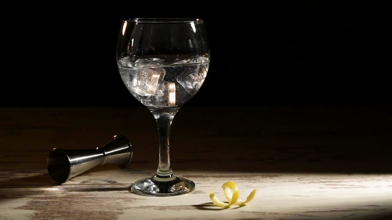persona vertiendo cubitos de hielo en un vaso de gin tonic con limón y jigger en la mesa en una habitación oscura