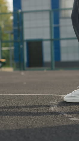 leg view of two women jogging on sports court, one in green joggers and one in ash joggers with white sneakers, blurred background featuring bar fence and people seated outdoors