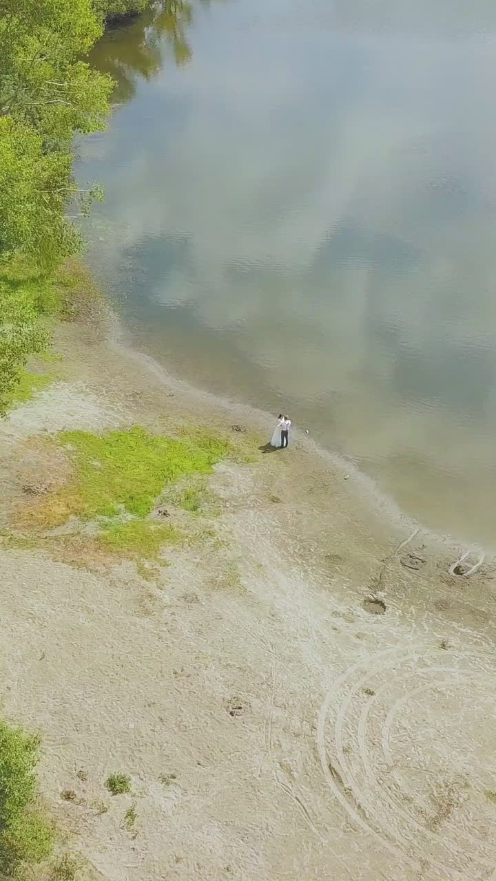 distant newlywed couple on green forestry bank of lake with mirror surface reflecting clouds bird eye view
