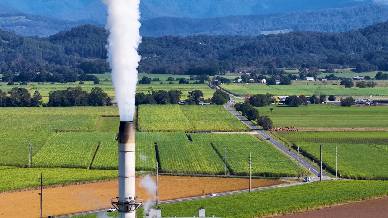 A factory chimney emits smoke over lush agricultural fields under clear skies, highlighting industrial impact on rural environments
