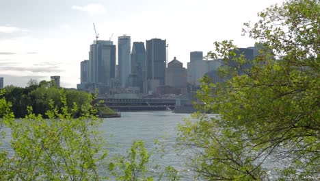 Downtown-Montreal-Skyline-From-Across-the-St-Lawrence-River-on-a-Sunny,-Hazy-Afternoon-with-Trees-and-Bushes-in-the-Foreground