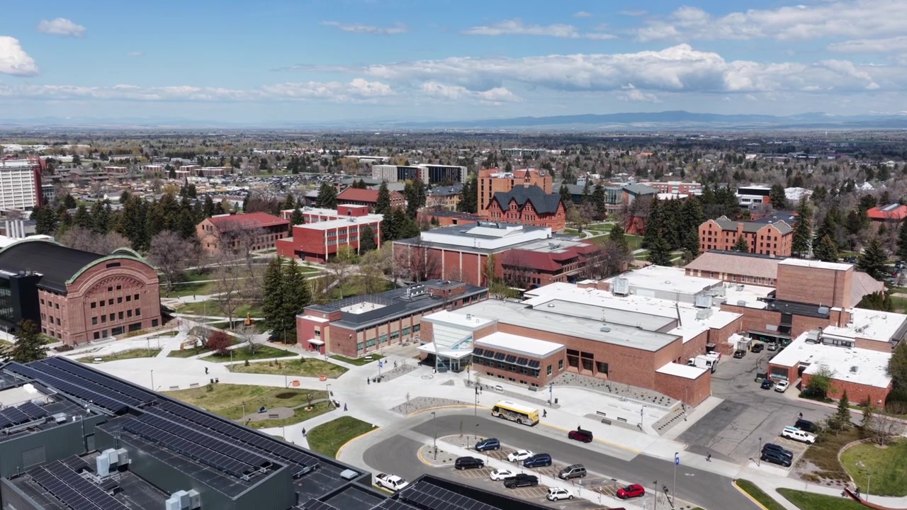 Montana State University-campus sprer seg over Bozeman, med akademiske mursteinsbygninger og solcellepaneler på taket, omgitt av grønne trær under fjerne, disige blå fjell. Bredt droneopptak som senker seg fra luften.