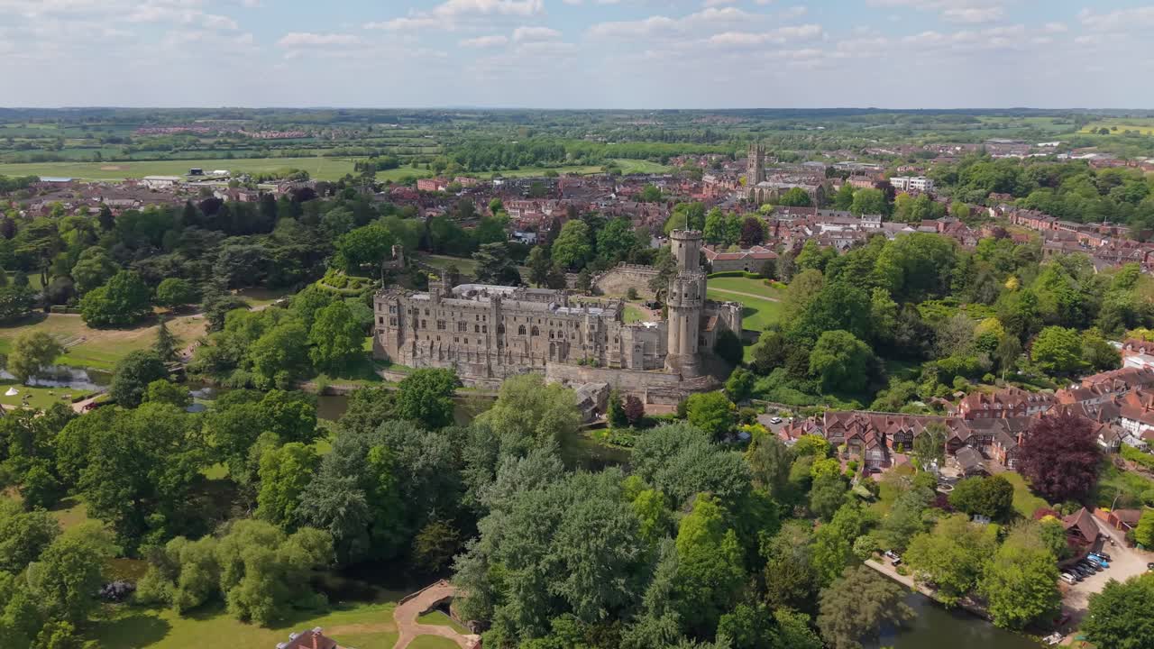Órbita de drone sobre a vila de Warwick na primavera, exibindo paisagens verdes exuberantes e o castelo histórico no centro, rodeada por uma paisagem rural encantadora e cenários sazonais vibrantes.