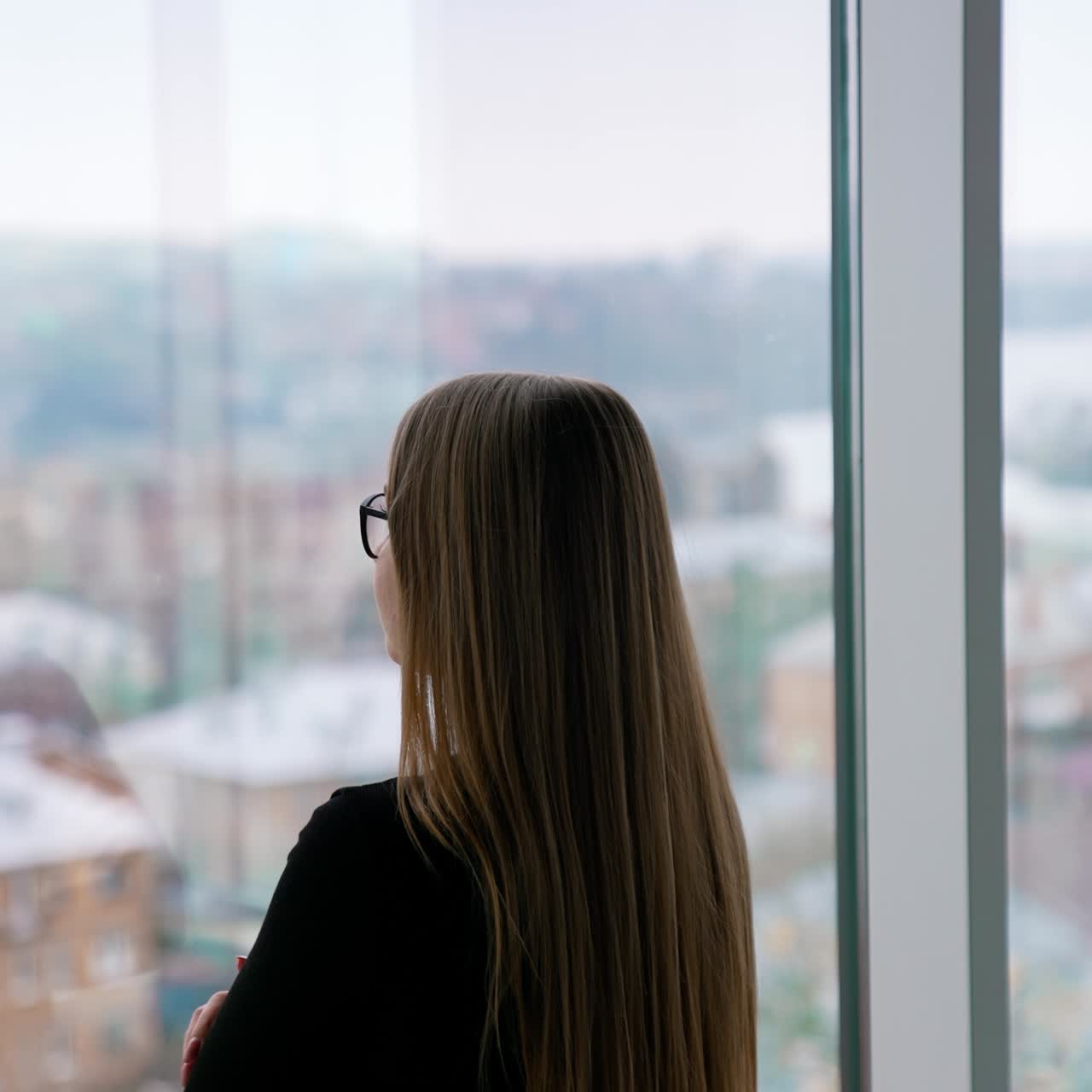 Attractive business lady in glasses standing at window. Long-haired woman looks through the window on a city landscape. Rear view