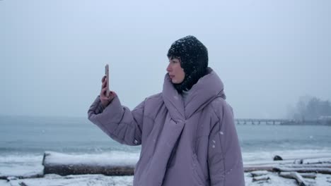 woman in warm clothing takes picture with smartphone on snowy beach