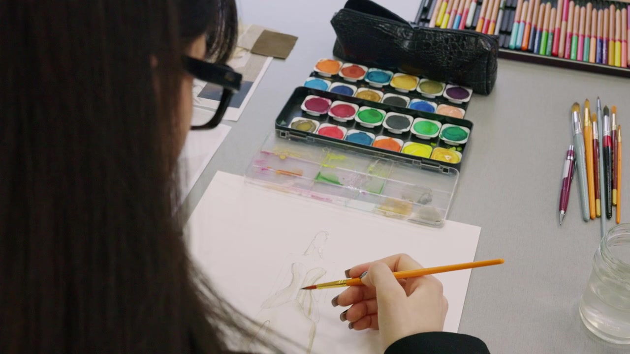 Close up of a hand painting a pale fashion figure sketch with an orange handled brush on white paper