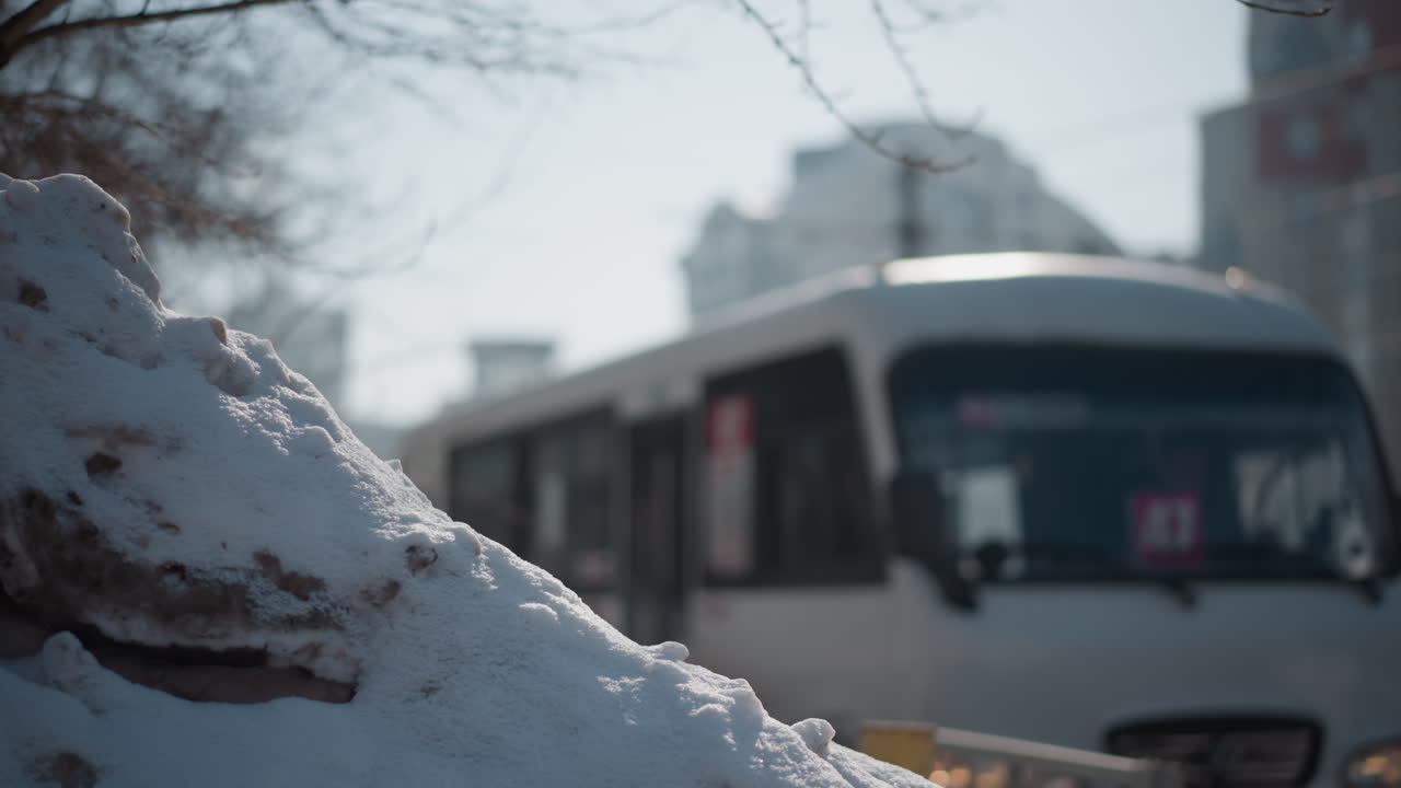 Snow heap beside road under bright winter sun, blurred buses glide along lane through city, headlights glint, soft bokeh on distant buildings, gritty plowed mound anchors foreground