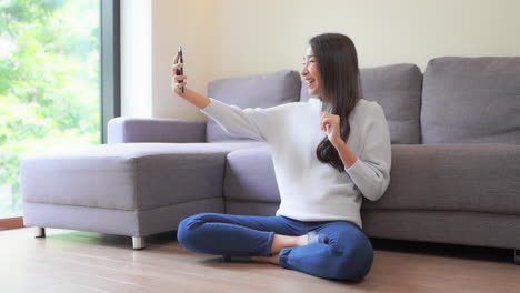 glad asian woman with a pleasant smile, waves with a hand at camera of cell phone in home interior, makes video call to her best friend sitting on a floor